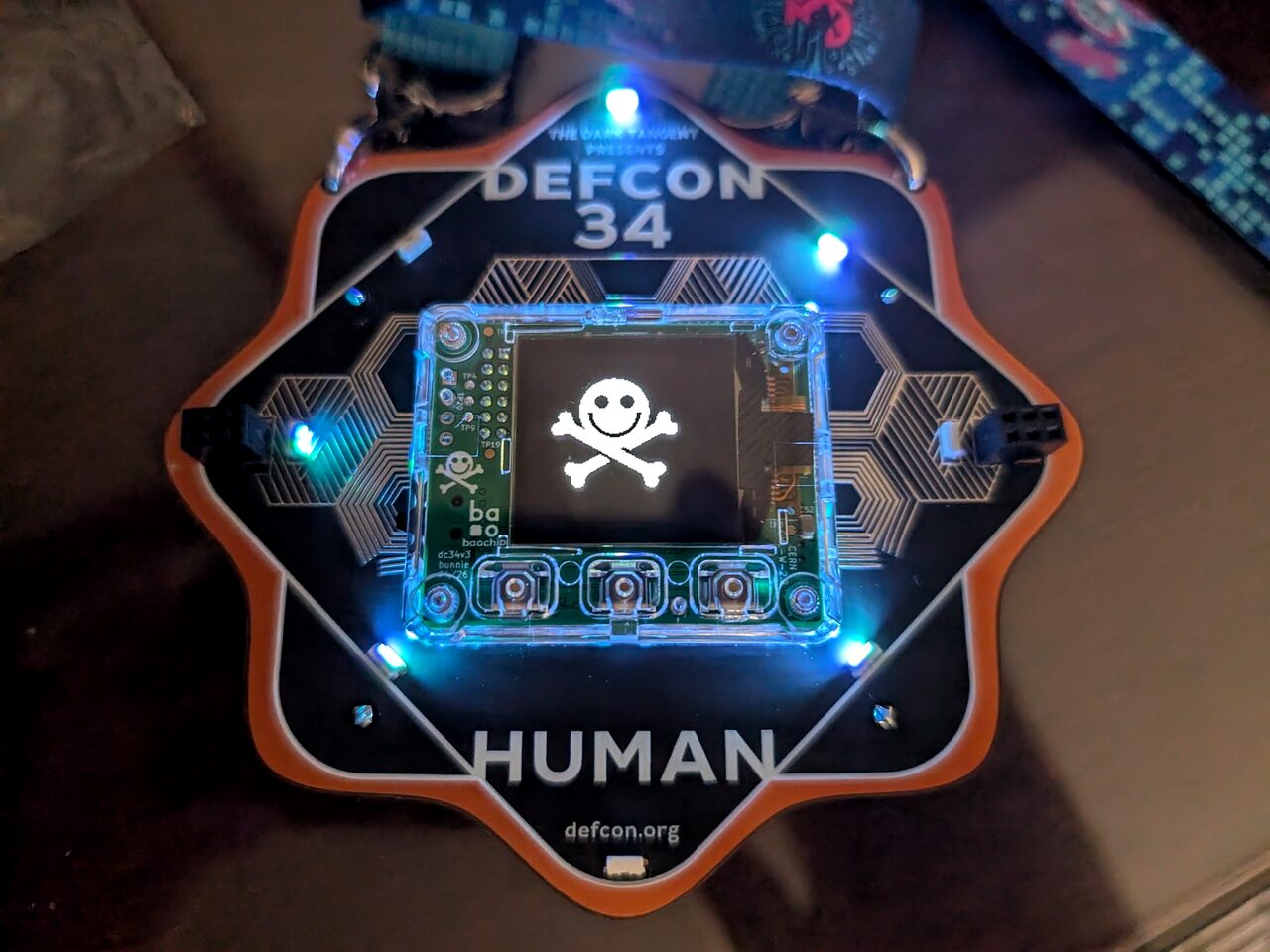 A lit DEF CON 34 Human badge showing a skull-and-crossbones on its monochrome screen.